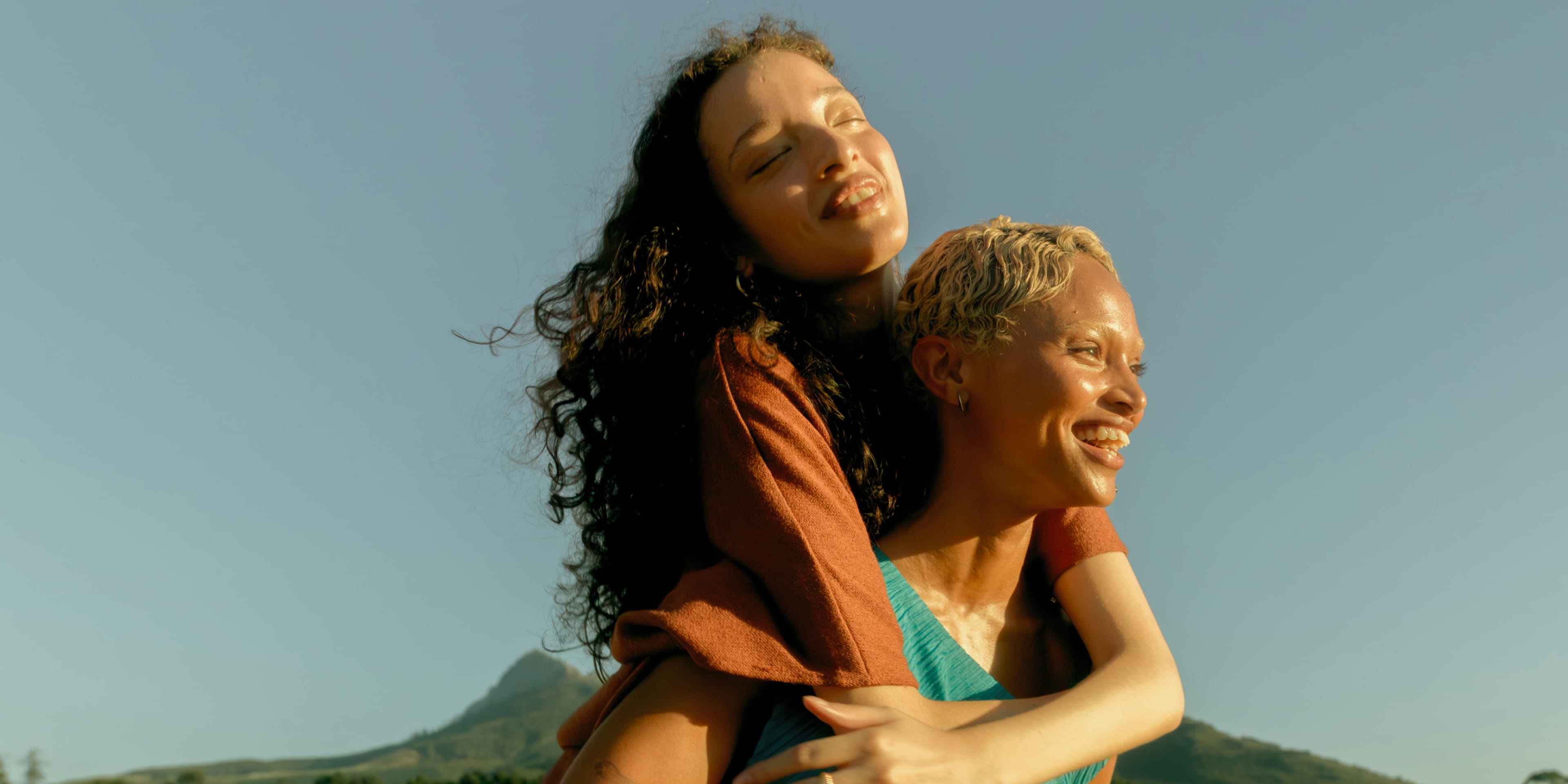 Two people laughing in a sunlit field, one giving the other a piggyback ride, with a mountain rising in the background.