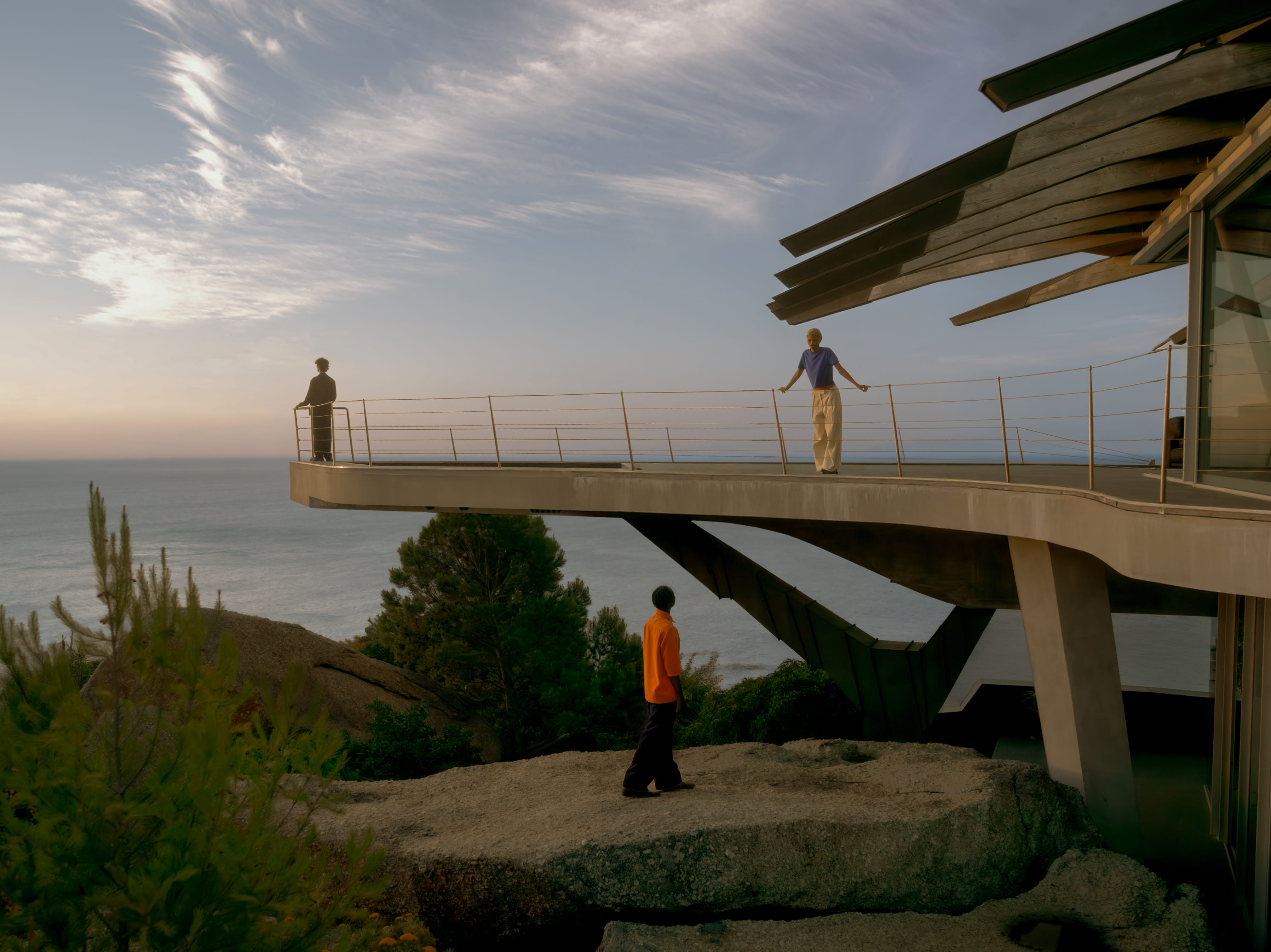 Three people occupy different levels of a modernist concrete building perched above the ocean at golden hour.