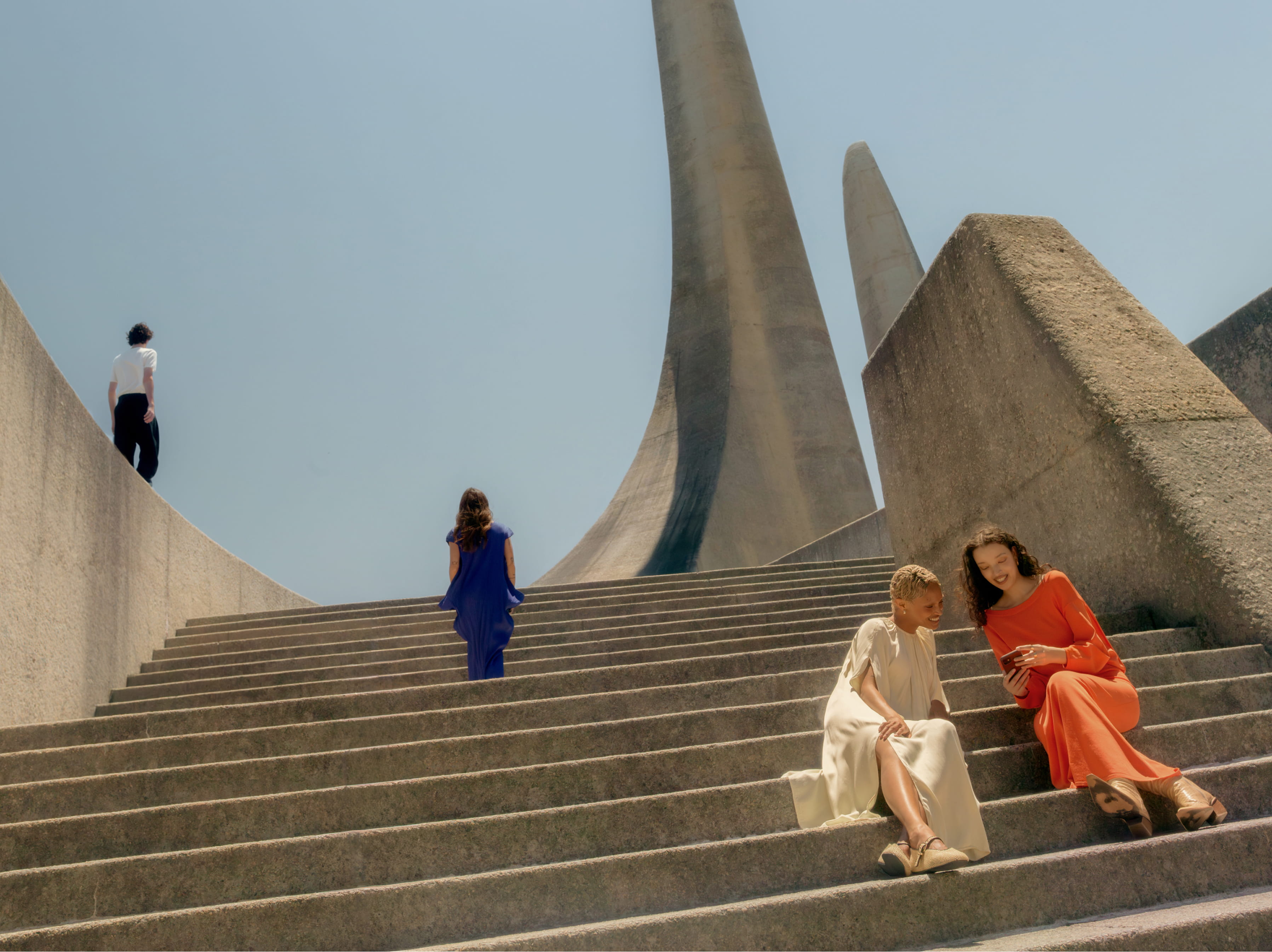 Two people sit together on wide concrete steps at a brutalist monument with towering pointed spires, smiling at something on a phone, while two other figures walk up the staircase behind them.