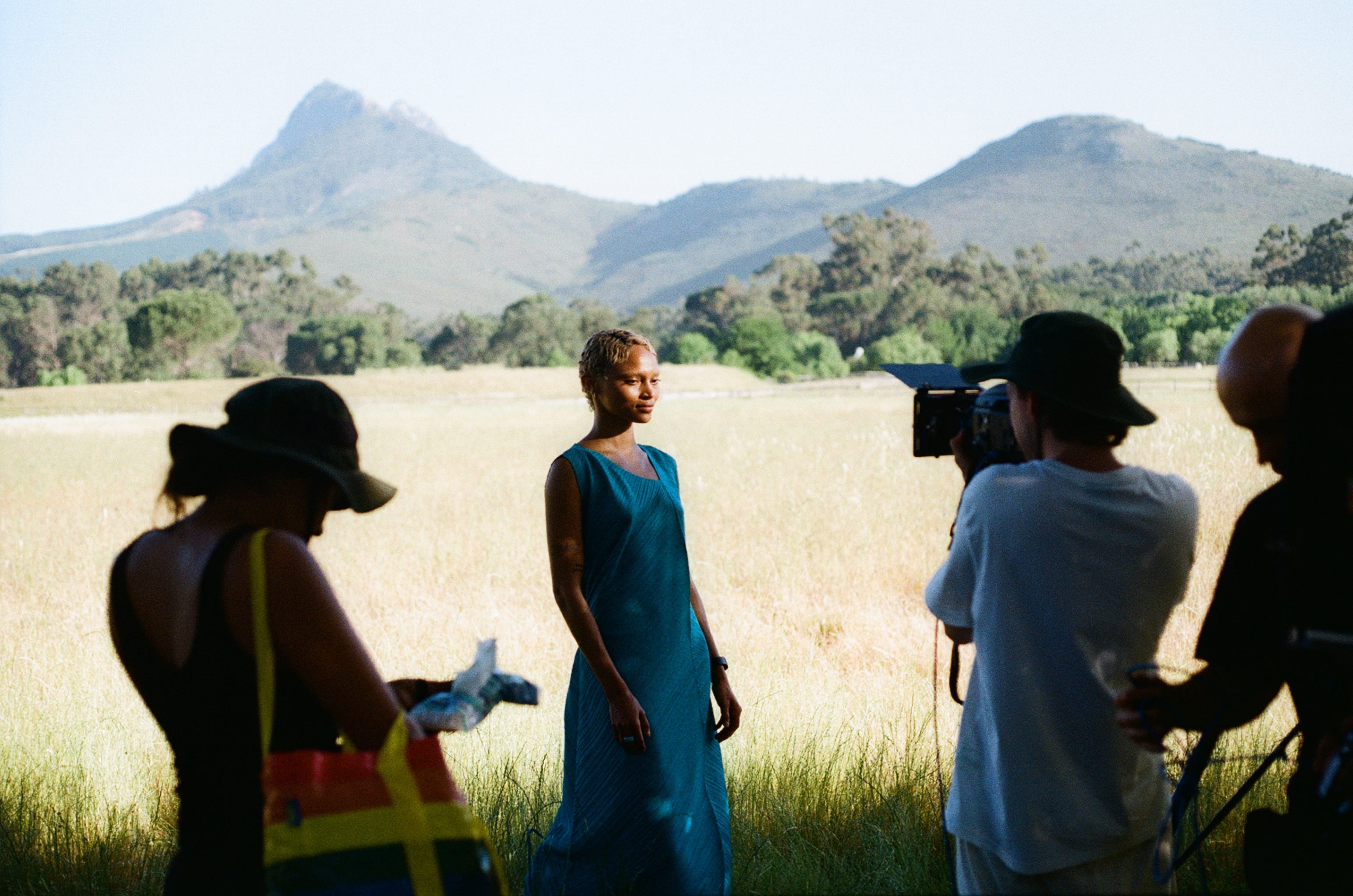 Behind-the-scenes shot of a model in a teal dress standing in a golden grass field with Cape Town mountains in the background, framed by silhouetted crew members with camera equipment.