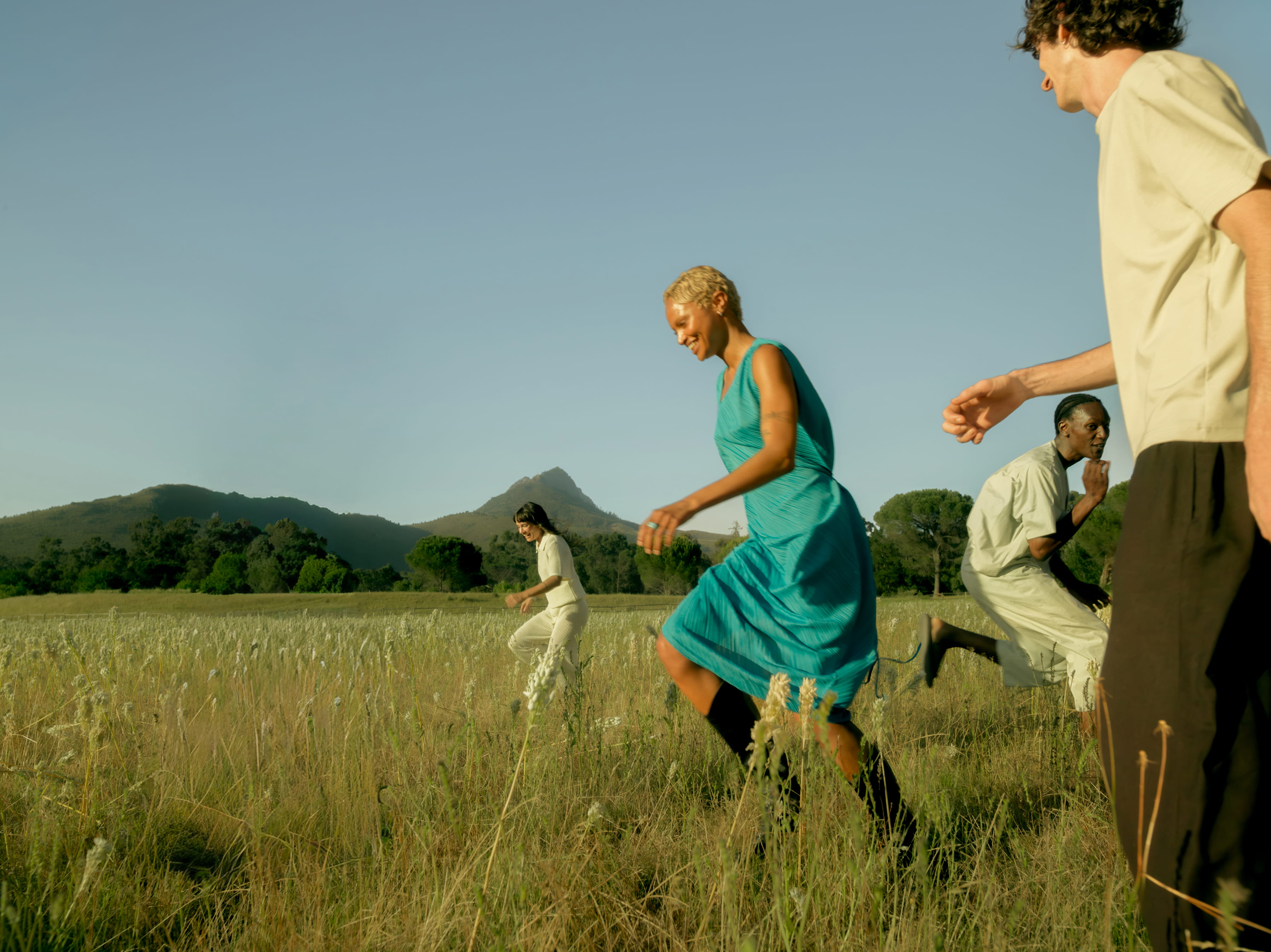 Four people stride through a field of tall grass and wildflowers with mountains behind them, captured mid-movement in warm, low-angled light.